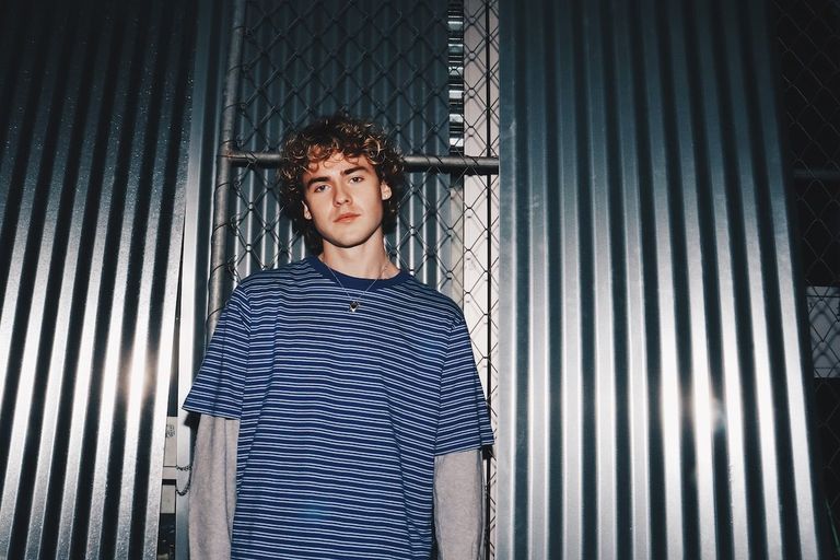 Musician Jack Gray in a blue striped shirt posing in front of corrugated metal walls and a chain-link fence. Photo by Grace Wethor.
